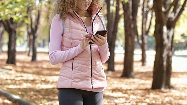 女人用智能手机发短信聊天户外散步，卷头发的女孩早晨秋天公园里金黄的树叶树视频下载