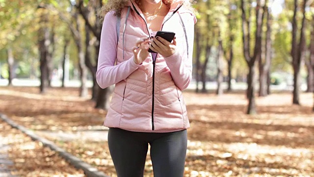 女人用智能手机发短信聊天户外散步，卷头发的女孩早晨秋天公园里金黄的树叶树视频素材