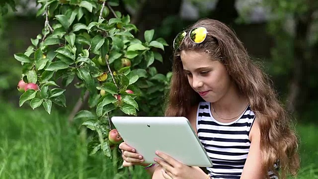 在花园里的苹果树下，戴着墨镜的漂亮女孩站在平板电脑上视频素材