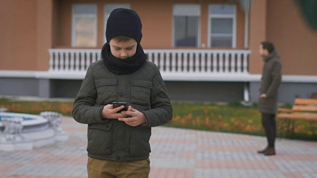 男孩穿着温暖的深灰色夹克，戴着黑色的帽子和围巾站在街上打电话，他的父亲在他身后。丈夫和儿子在等他们的妻子和母亲视频素材