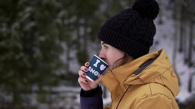 戴着黑色泡泡帽的年轻女子在雪地里走到树旁，从纸杯里喝着热饮料视频素材