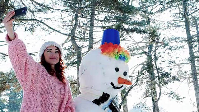 冬季自拍，可爱的女孩与雪人合影，一个年轻女子在手中的手机在冬季森林背光做有趣的自拍照片视频素材