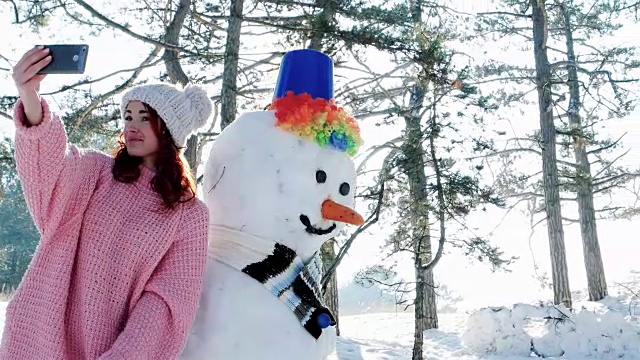 可爱的女孩与雪人合影，冬天的自拍，一个年轻的女人在冬天森林背光做有趣的自拍照片在手手机视频素材