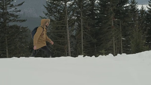 一个穿着棕色夹克、带着兜帽、背着背包的人穿过了冬天下雪的森林视频素材