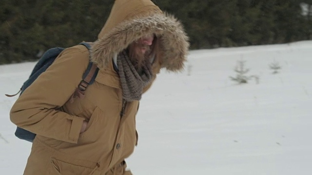 一个穿着棕色夹克、带着兜帽、背着背包的人穿过了冬天下雪的森林视频素材