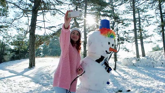 手机在一个年轻的女人在冬季森林背光自拍照片，可爱的女孩与雪人合影视频素材