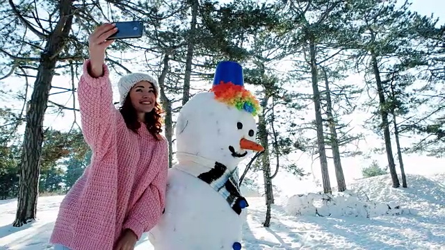 可爱的女孩与雪人合影，冬天的自拍，一个年轻的女人在冬天森林背光做有趣的自拍照片在手手机视频素材