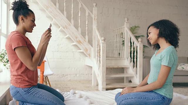 一名快活的混血女子坐在家里卧室的床上用智能手机拍摄他的女朋友视频素材