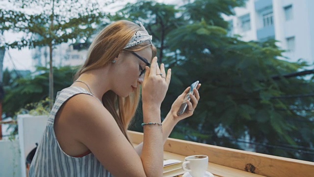 一个女人在咖啡馆里用智能手机上的应用程序。美丽的年轻女孩微笑着摘下她的眼镜。视频素材