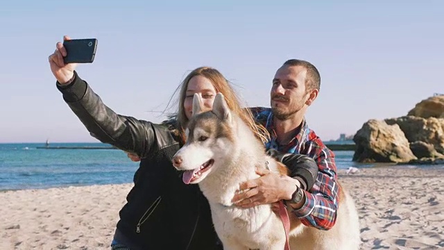 一对年轻的白人夫妇在海滩上用智能手机和西伯利亚哈士奇狗自拍，慢动作视频素材