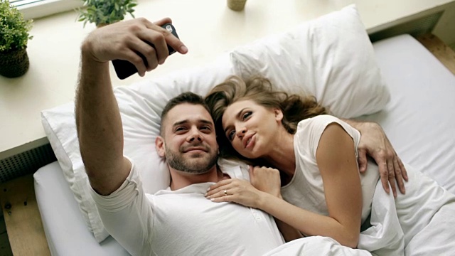 年轻美丽的情侣用智能手机自拍，早上躺在床上接吻视频素材
