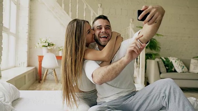 年轻美丽的恩爱夫妻用智能手机自拍，早上坐在床上亲吻视频素材