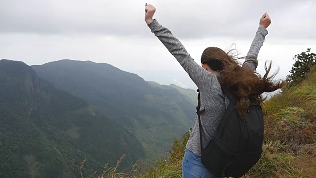 一名年轻女性徒步旅行者背着背包爬上山顶，举起双手。一名女游客站在美丽的峡谷边缘，胜利地伸出双臂。慢镜头后视图视频素材