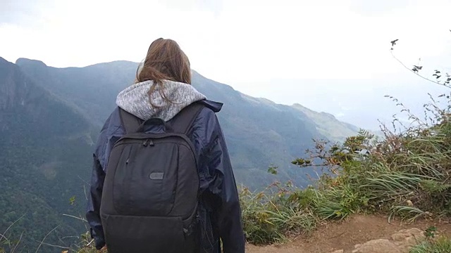 一名年轻女性徒步旅行者背着背包爬上山顶，举起双手。一名穿着雨衣的女游客站在美丽的峡谷边缘，胜利地伸出双臂。慢动作视频素材