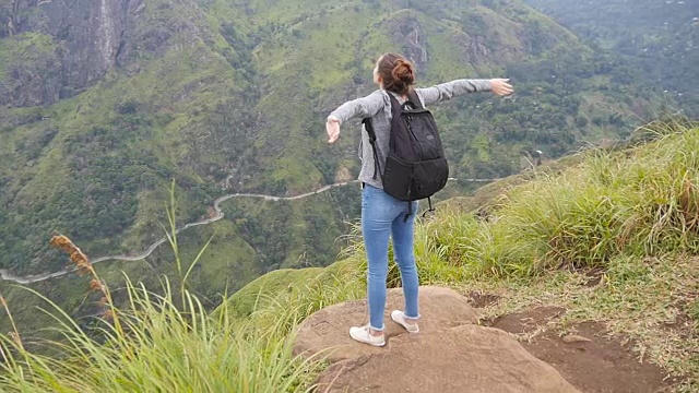 一名女游客站在美丽的峡谷边缘，胜利地伸出双臂。一名年轻女性徒步旅行者背着背包爬上山顶，举起双手。慢镜头后视图视频素材