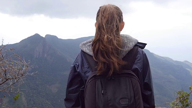 陌生的女游客站在美丽的峡谷边缘欣赏风景。年轻的女徒步旅行者穿着雨衣和背包到达山顶，举起手。后方后视图视频素材