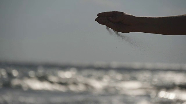 日落时分，在大海的背景下，女人们用手把海沙从手指间倒出来。年轻女子的手臂上撒满了沙子。沙砾从女孩的拳头。慢镜头视频素材