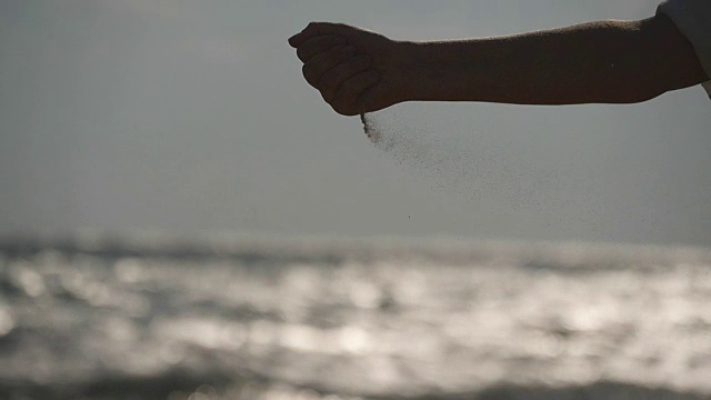 日落时分，在大海的背景下，女人们用手把海沙从手指间倒出来。年轻女子的手臂上撒满了沙子。沙砾从女孩的拳头。慢镜头视频素材