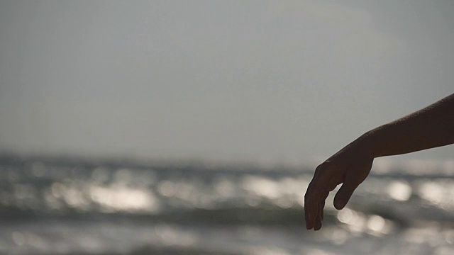 日落时分，在大海的背景下，女人们用手把海沙从手指间倒出来。年轻女子的手臂上撒满了沙子。沙砾从女孩的拳头。慢镜头视频素材