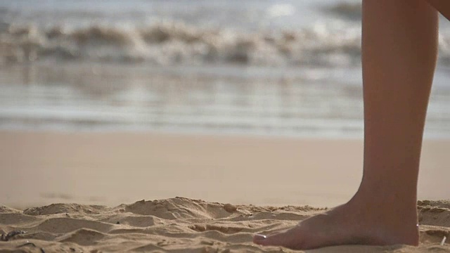 近距离的女性的脚走在金色的沙滩与海浪的背景。年轻女子的双腿踩在沙子上。光着脚的女孩在海边。暑假假期。慢动作视频素材