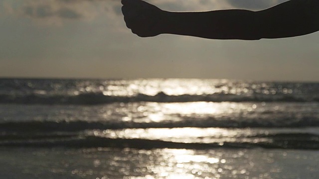 日落时分，在大海的背景下，女人们用手把海沙从手指间倒出来。年轻女子的手臂上撒满了沙子。沙砾从女孩的拳头。慢镜头视频素材