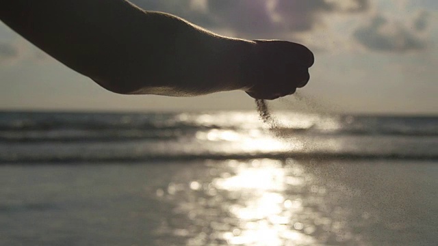 日落时分，在大海的背景下，女人们用手把海沙从手指间倒出来。年轻女子的手臂上撒满了沙子。沙砾从女孩的拳头。慢镜头视频素材