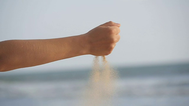 女人的手通过她的手指倒海沙在海洋的背景。年轻女子的手臂上撒满了沙子。沙砾从女孩的拳头。慢镜头视频素材