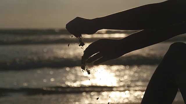 玩沙子的年轻女子的手臂。日落时分，在大海的背景下，女人们用手把海沙从手指间倒出来。慢镜头视频素材