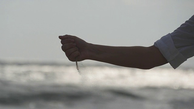 日落时分，在大海的背景下，女人们用手把海沙从手指间倒出来。年轻女子的手臂上撒满了沙子。沙砾从女孩的拳头。近距离视频素材