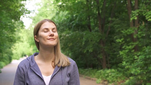 快乐微笑的年轻女子穿着帽衫漫步在阳光明媚的公园在夏天的一天视频素材