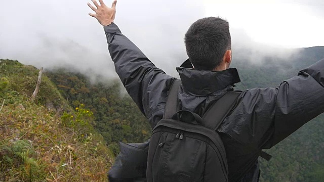 一名年轻男性徒步旅行者背着背包爬上山顶，举起双手。一名游客站在美丽的峡谷边缘，胜利地伸出双臂。慢镜头后视图视频素材