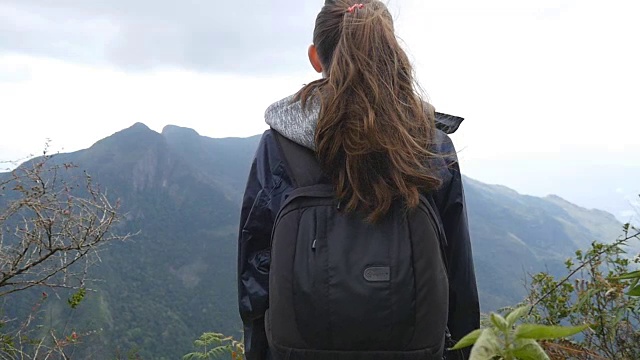 年轻的女游客背包欣赏美丽的风景在山。穿着雨衣的女旅行者站在美丽的峡谷边缘，欣赏大自然。慢镜头后视图视频素材