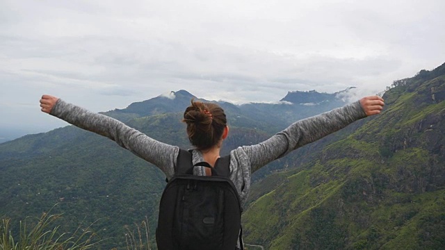 一名年轻女性徒步旅行者背着背包爬上山顶，举起双手。一名女游客站在美丽的峡谷边缘，胜利地伸出双臂。慢镜头后视图视频素材