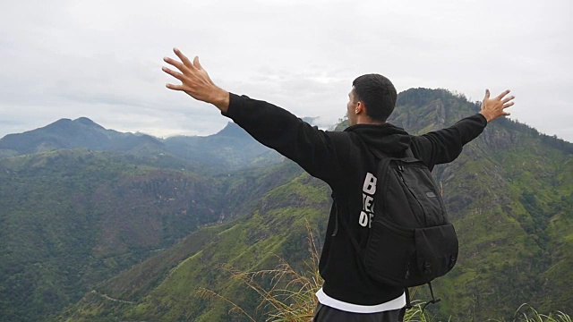 一名年轻男性徒步旅行者背着背包爬上山顶，举起双手。一名游客站在美丽的峡谷边缘，胜利地伸出双臂。慢镜头后视图视频素材