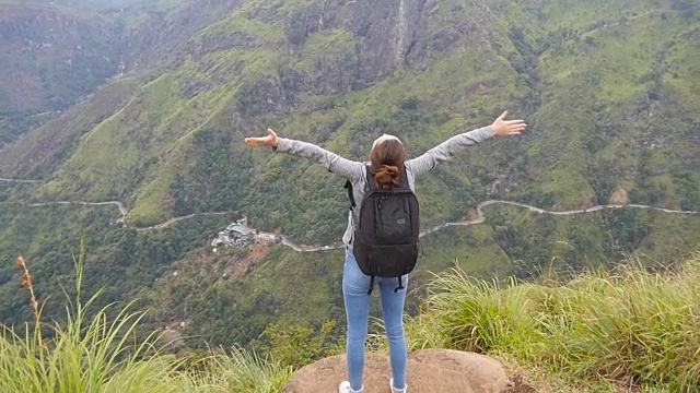 一名年轻女性徒步旅行者背着背包爬上山顶，举起双手。一名女游客站在美丽的峡谷边缘，胜利地伸出双臂。慢镜头后视图视频素材