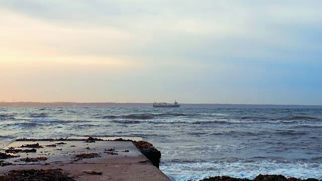 日落时海浪撞击码头的慢镜头，远处的货船在背景中视频素材