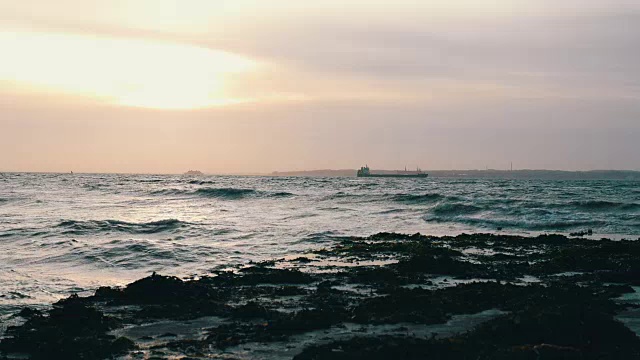 黄昏时分，海浪拍打着码头，远处的货船在背景中慢镜头地出现视频素材