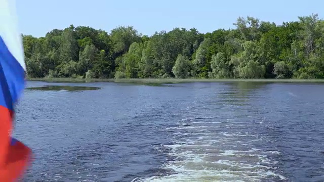 船尾挂着一面俄罗斯国旗。波浪和在水面上醒来。夏天的河流或湖泊。蓝色的天空。河岸上长满了绿色的植被。船马达的声音。视频剪辑4K，超高清视频素材