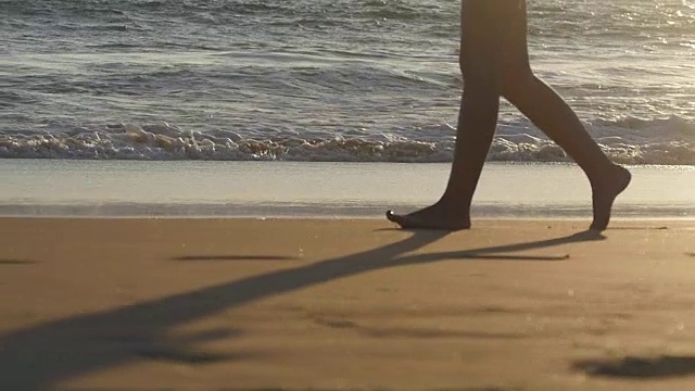 近距离的女性的脚走在金色的沙滩与海浪的背景。年轻女子的双腿踩在沙子上。光着脚的女孩在海边。暑假假期。慢动作视频素材