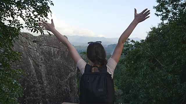 一名年轻女性徒步旅行者背着背包爬上山顶，举起双手。一名女游客站在美丽的峡谷边缘，胜利地伸出双臂。慢镜头后视图视频素材