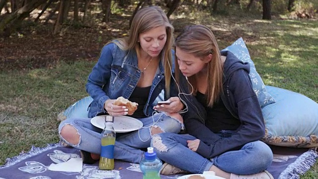 两个年轻漂亮的女孩一边在手机上听音乐，一边在公园里吃午饭视频素材