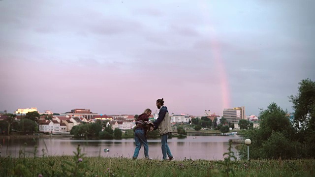 美丽的年轻男子和女子站在河岸上，在夕阳下拥抱，背景是彩虹视频素材