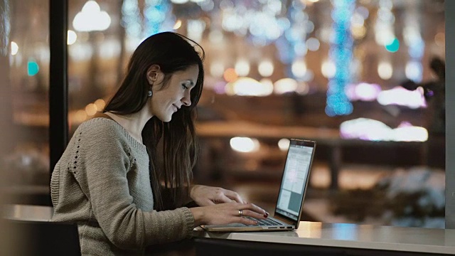 年轻漂亮的女人晚上坐在窗边，用笔记本电脑工作。女孩上网视频素材