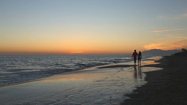 一对年轻夫妇在夕阳下沿着海滩散步，远离镜头视频素材