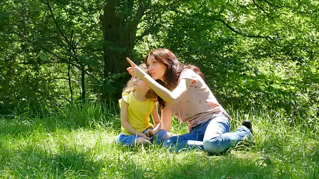 有吸引力的年轻母亲和小女儿的肖像花时间一起坐在绿色的草地在城市公园和谈话。视频素材