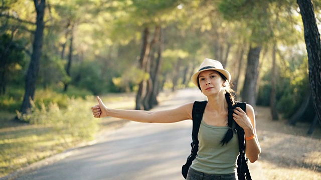 旅行者女人搭便车在一个阳光明媚的森林道路。旅游女孩找车开始她的旅程视频素材