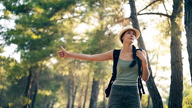 旅行者女人搭便车在一个阳光明媚的森林道路。旅游女孩找车开始她的旅程视频素材