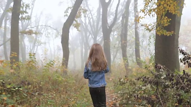 一个长头发的欧洲女孩，蓝色的夹克，黑色的裤子，运动鞋和蓝色的眼睛。一个抱着的小孩正跑着穿过雾蒙蒙的荒凉的森林。孤独。视频素材