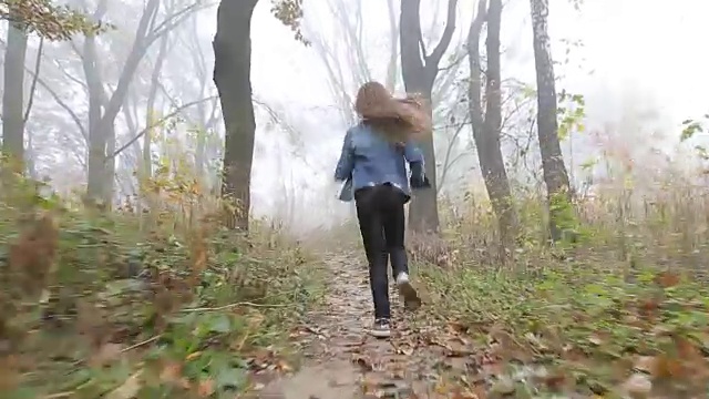 一个长头发的欧洲女孩，蓝色的夹克，黑色的裤子，运动鞋和蓝色的眼睛。一个吓坏了的小孩在雾蒙蒙的荒凉的森林里跑着。孤独。视频素材