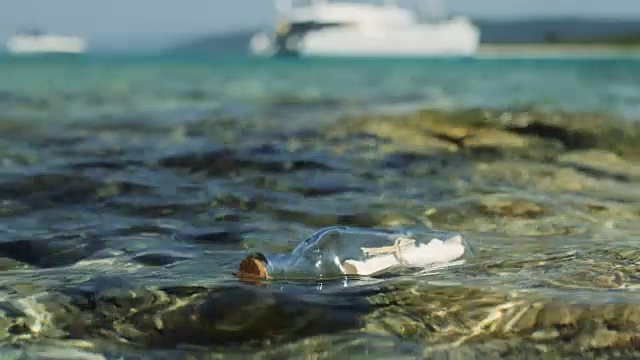 一个瓶子里的信息的特写镜头，漂流在浅海与白色游艇在背景。视频素材